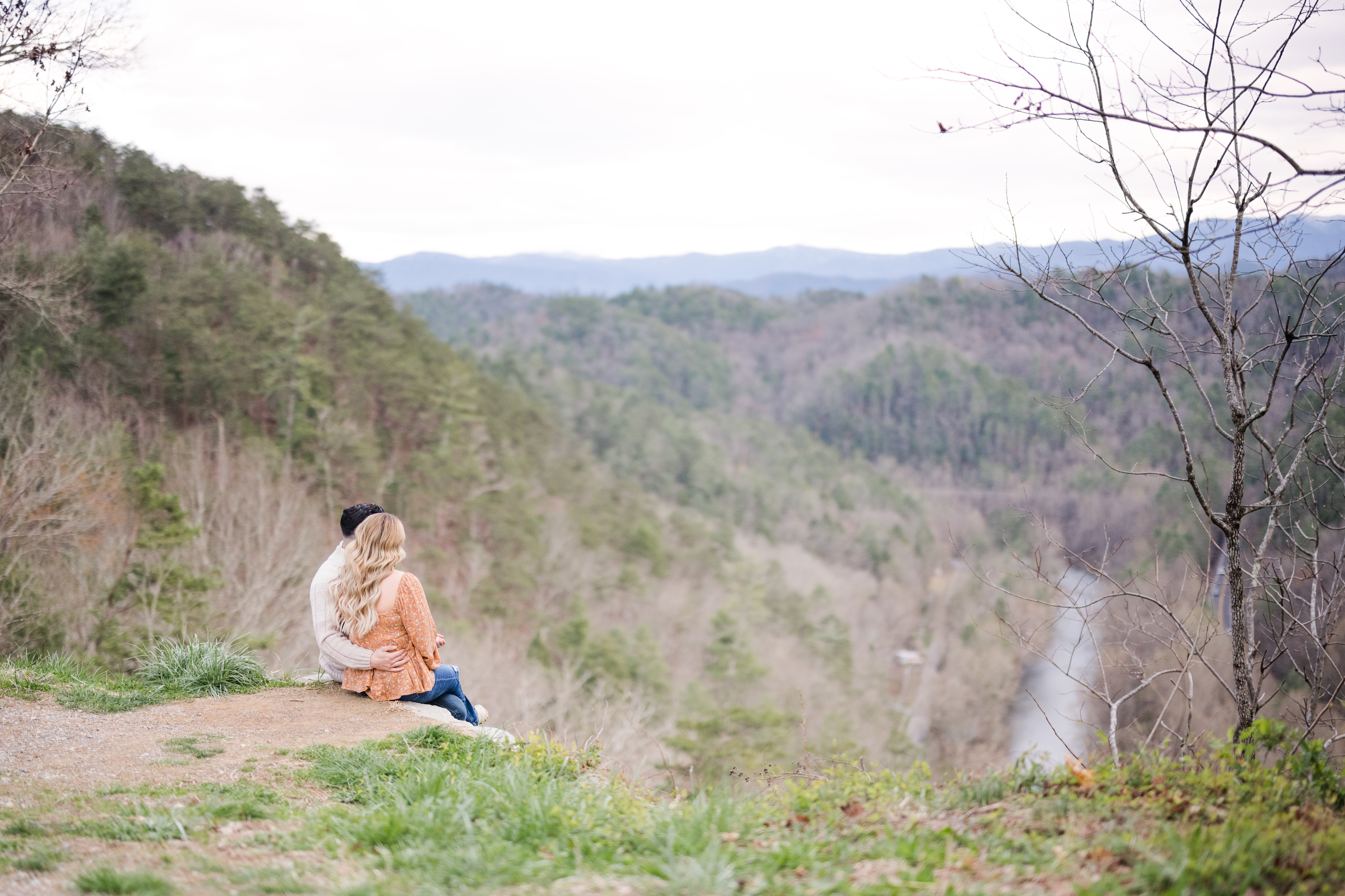 Gatlinburg engagement photographer captures couple snuggled at the edge of a cliff overlooking mountains and river during Smoky Mountains engagement session.