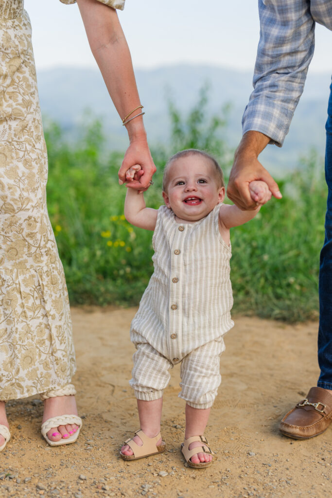 a mom and a dad each take their infants hand to help him stand up as he is smile stretches across his face with a view of the great smoky mountains national park behind them