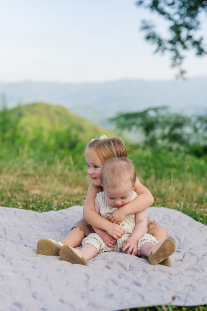A little girl sitting on a blanket hugging her baby brother with the Smoky Mountains in the background