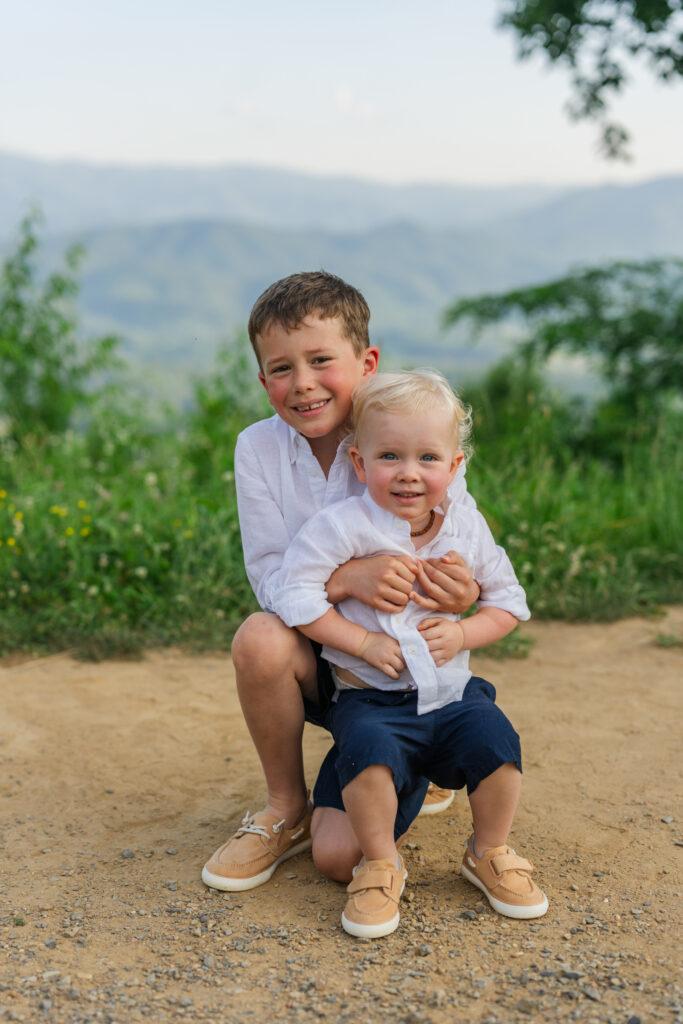 a big brother hugs his little brother from behind as they both are grinning ear to ear while having their family portraits taken by a professional photographer in Gatlinburg, Tennessee