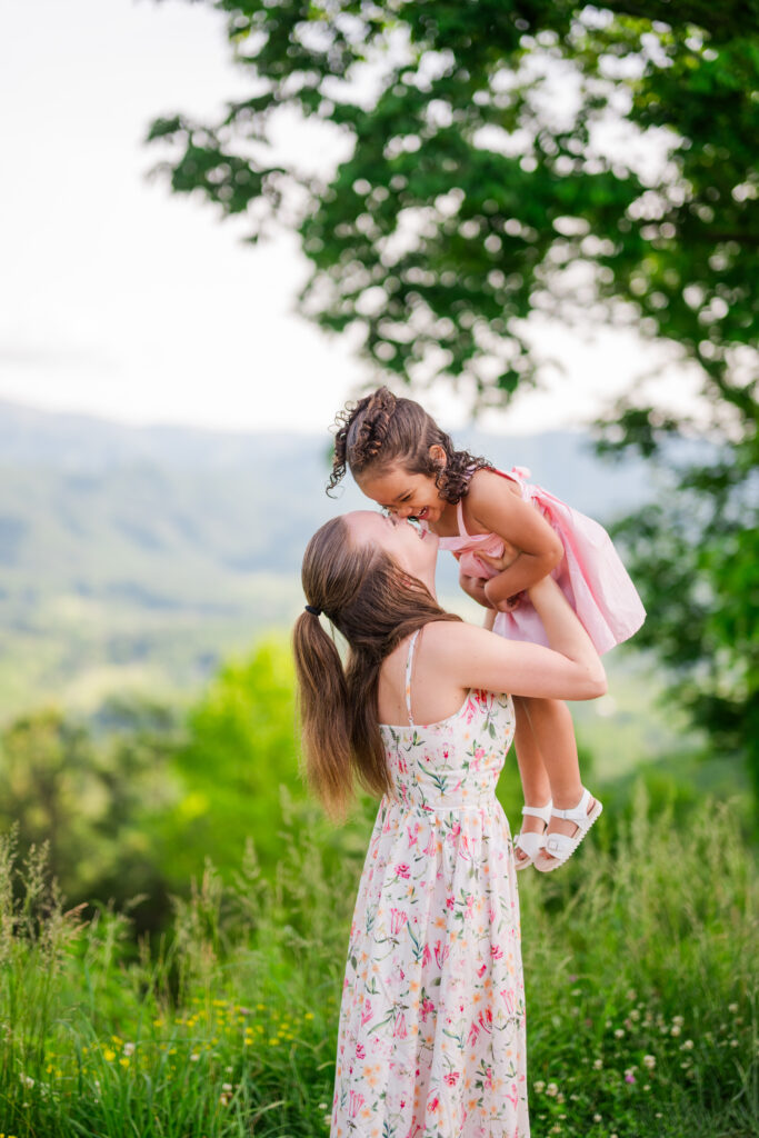 A mom lifting her young daughter above her head and bringing her back down for nose kisses on a beautiful day while on vacation in the smoky mountains in springtime with a view of the mountains and lush greenery