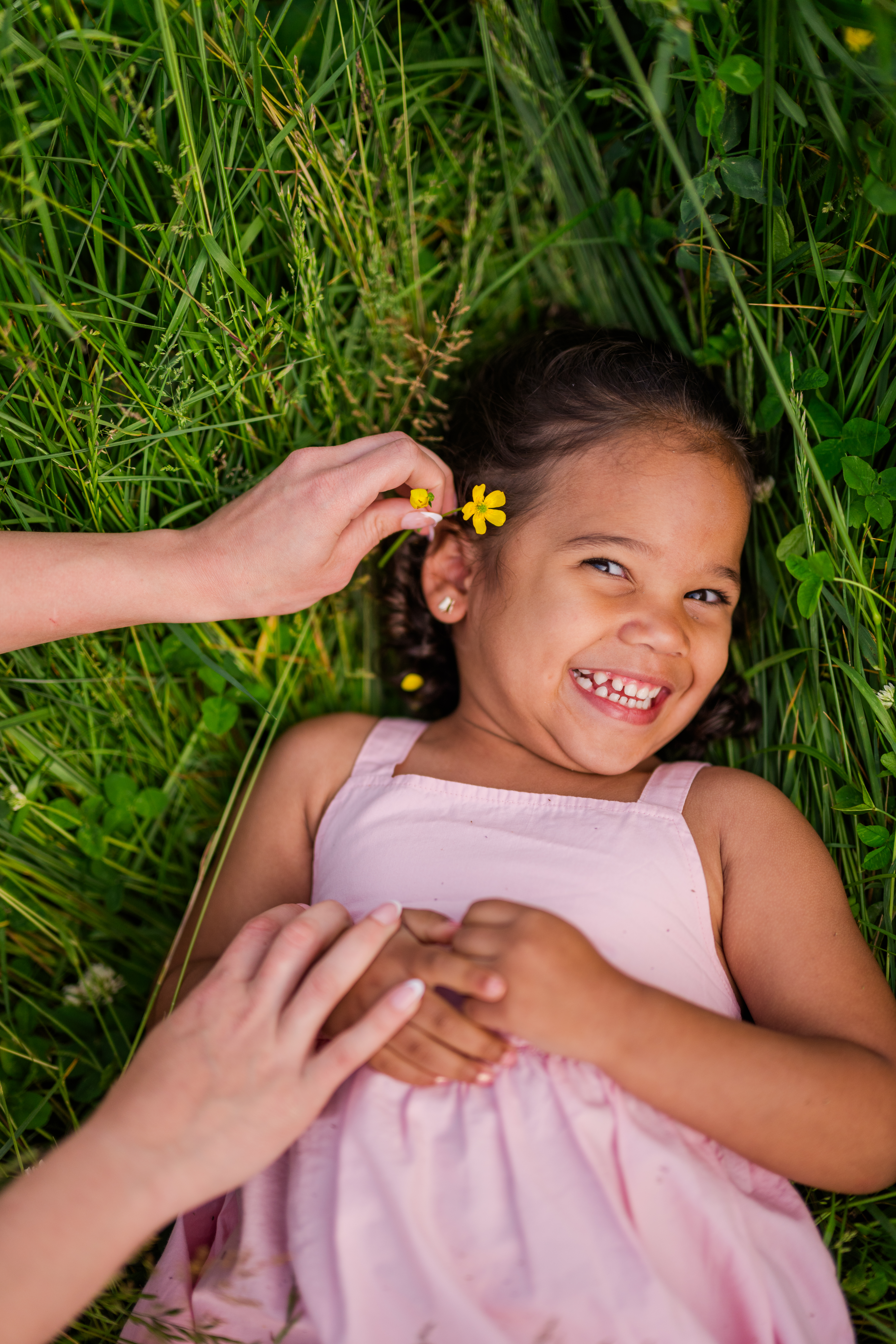 Pigeon Forge family photographer captures young girl smiling in tall spring grass as her mom places a yellow flower behind her ear during Smoky Mountains vacation.