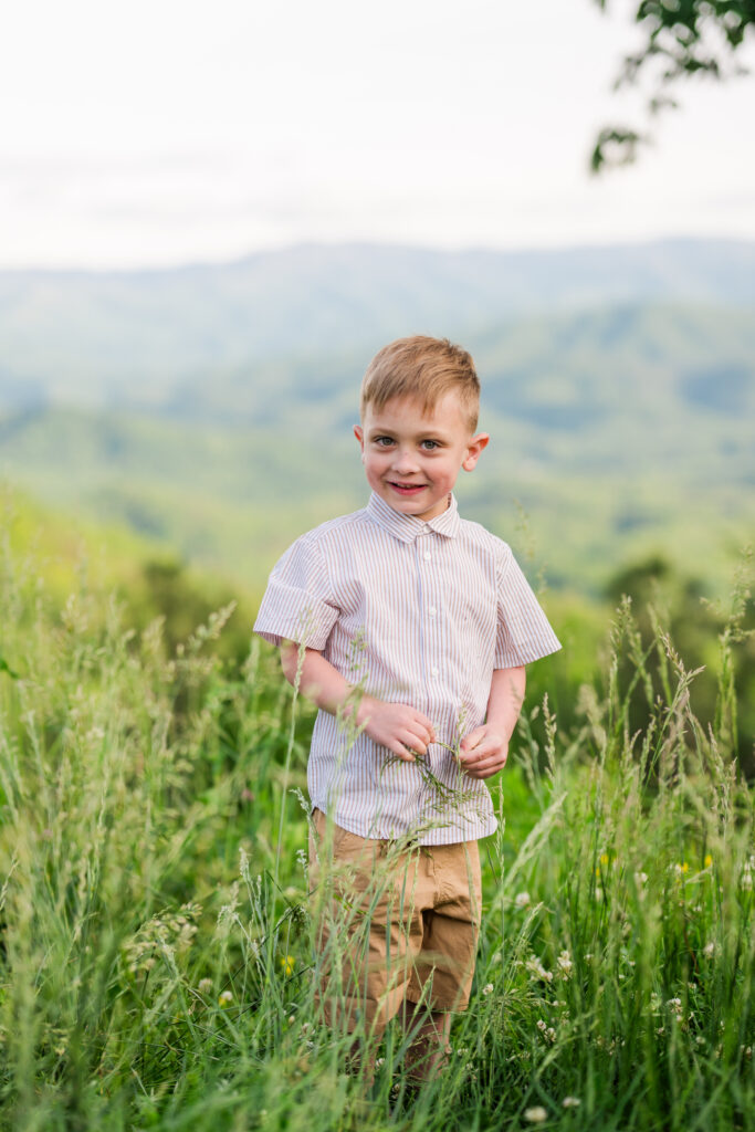 Smoky Mountain vacation photography of young boy smiling in tall grass with mountain views in Gatlinburg, Tennessee.