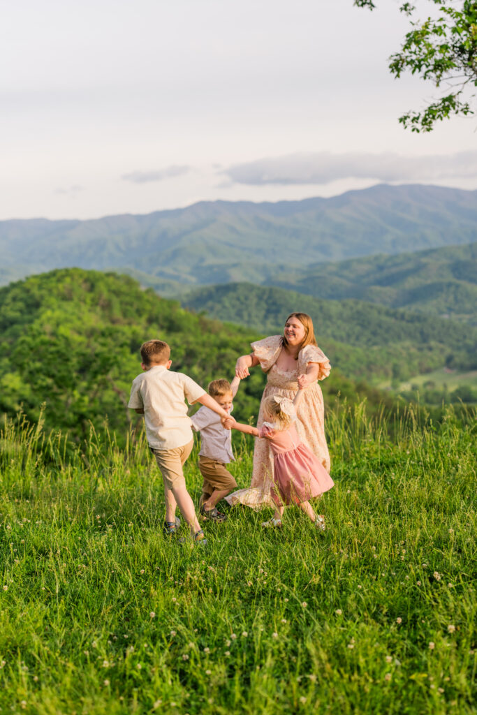 Smoky Mountain family photographer captures mother playing Ring Around the Rosie with her three kids at sunset overlooking the mountains in Great Smoky Mountains National Park.