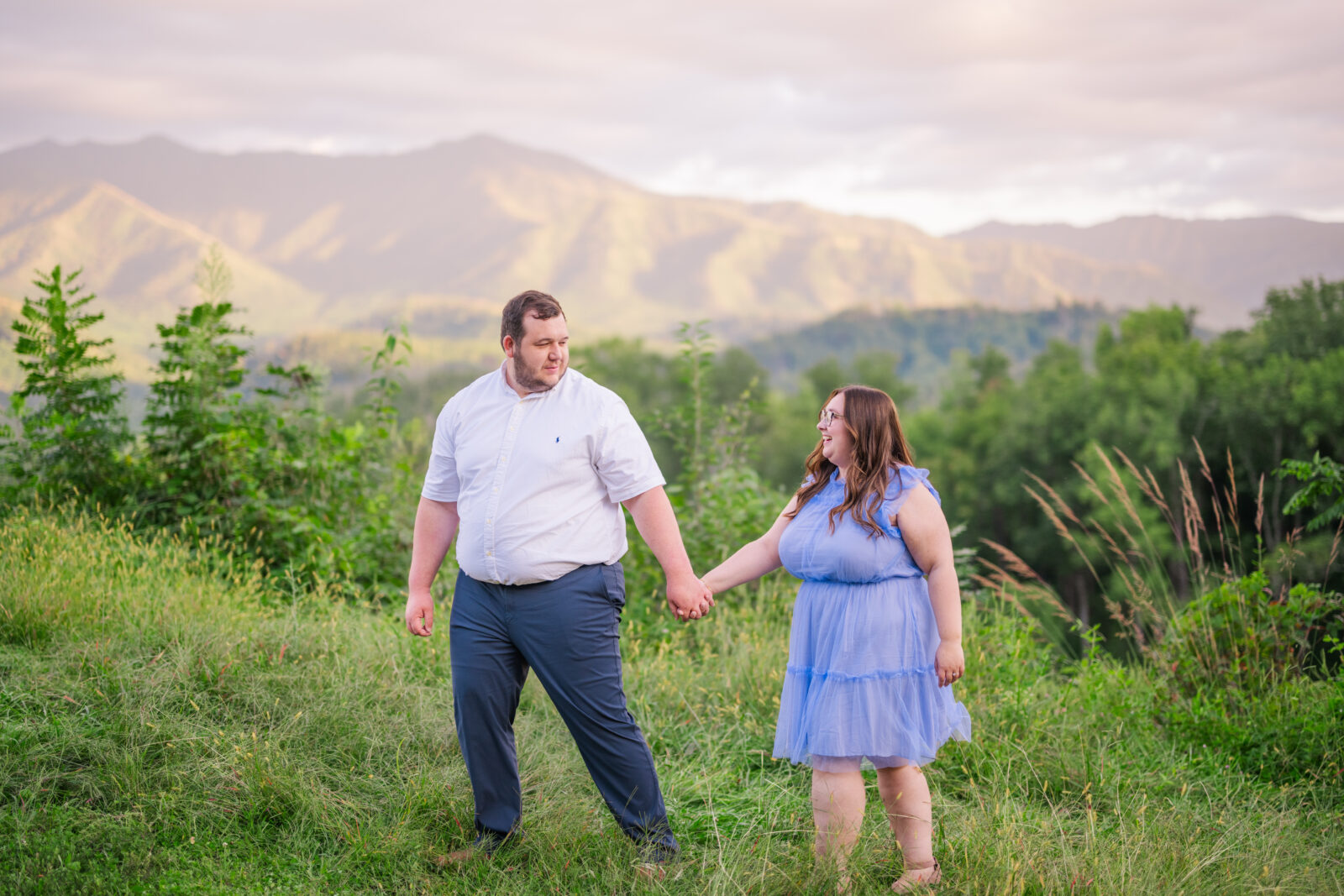 Photographer captures man leading his fiancé by the hand through a field at sunset during engagement session at Gatlinburg scenic overlook in Great Smoky Mountains National Park.