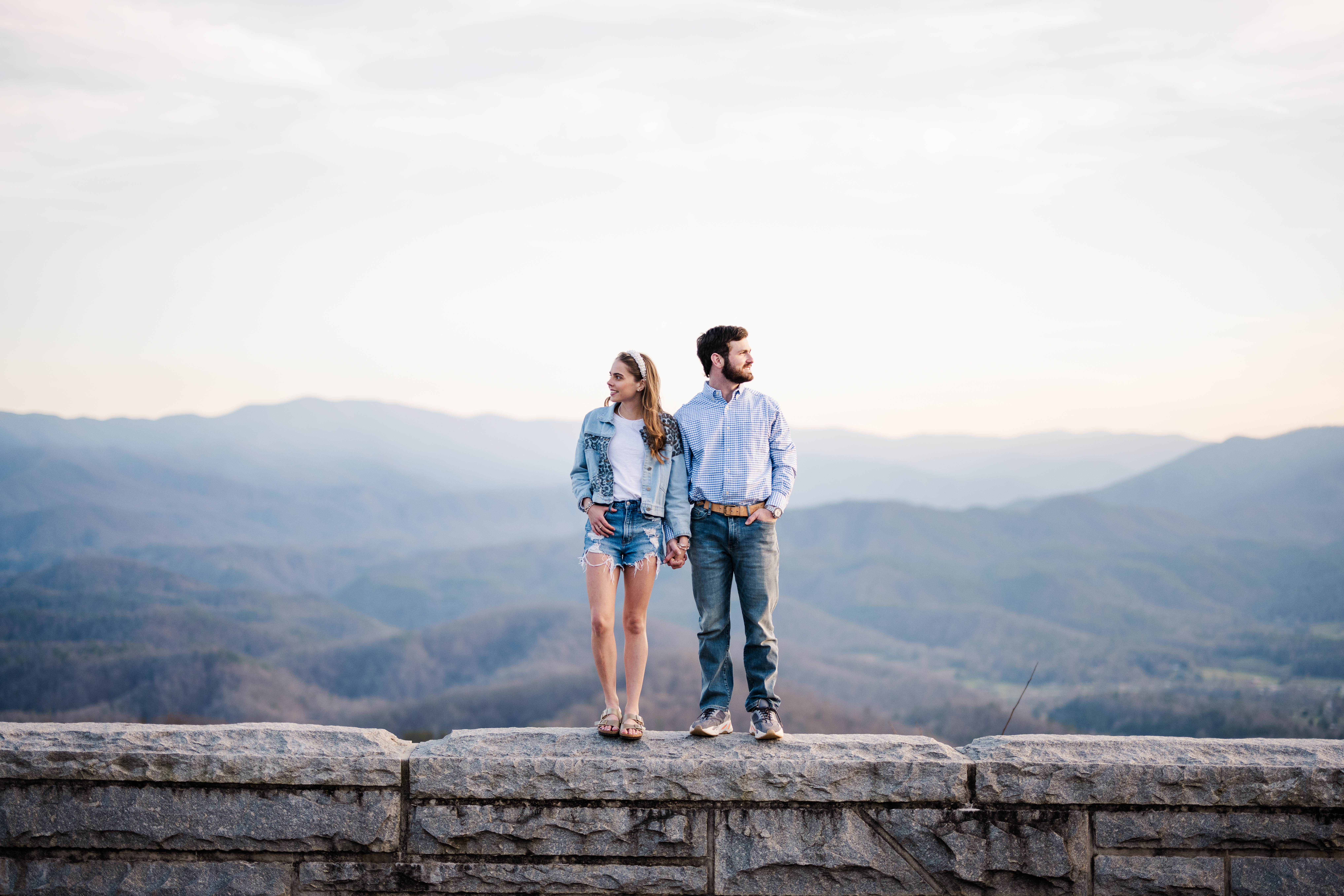 Gatlinburg photographer captures couple standing on rock wall holding hands with mountains in the background during engagement session at Foothills Parkway in Great Smoky Mountains National Park
