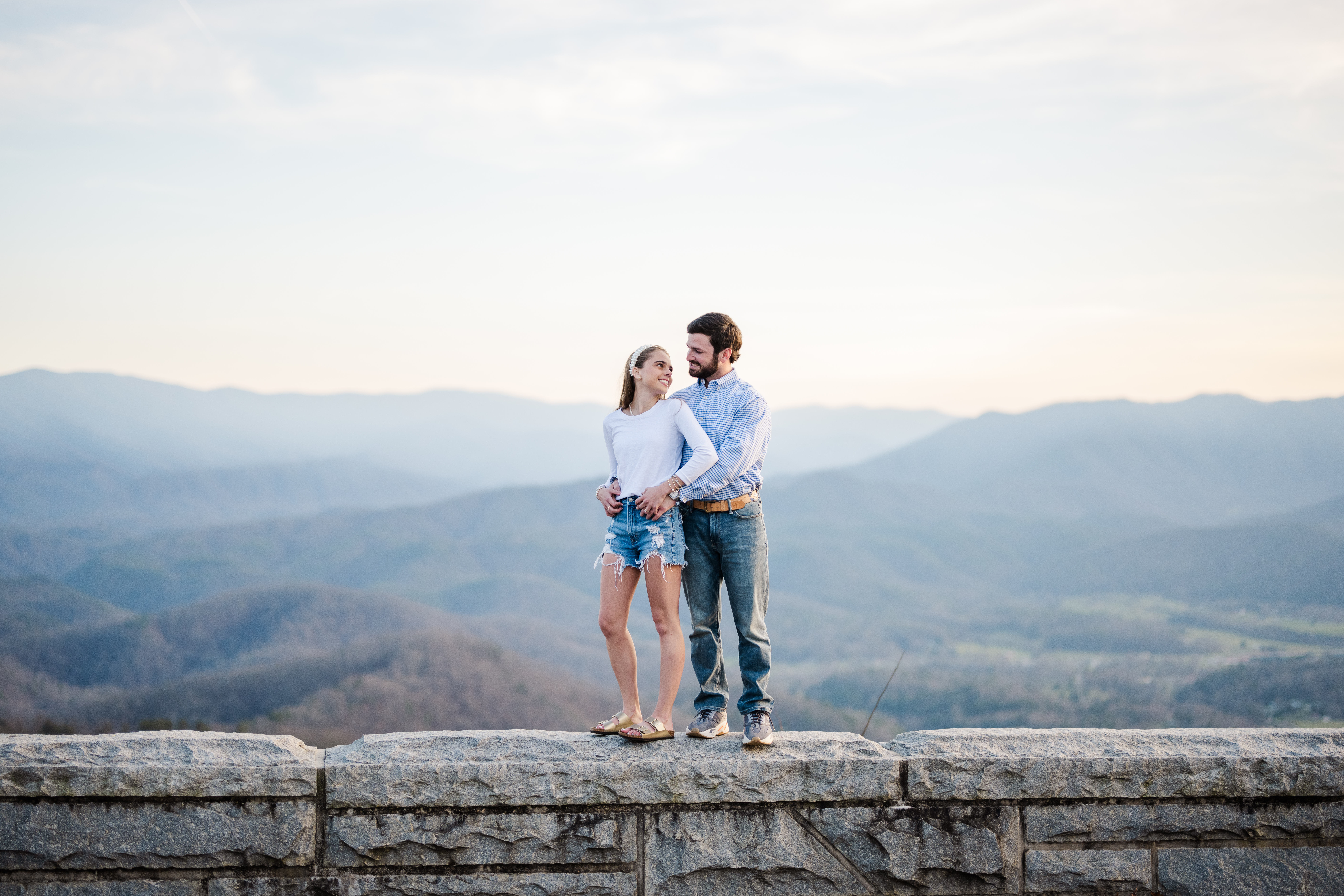 Gatlinburg engagement photographer captures couple holding each other on a rock wall with layered blue mountains in the background during Great Smoky Mountains National Park engagement session.