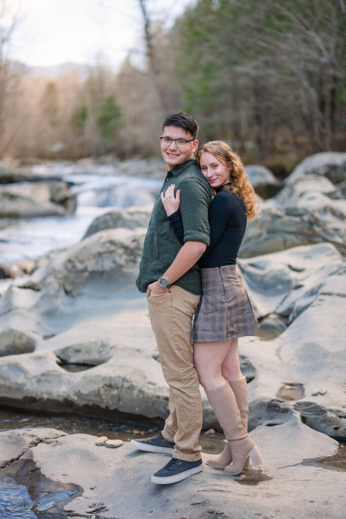 Engaged couple smiling as woman hugs man from behind by the river during engagement session in Great Smoky Mountains National Park while visiting Gatlinburg, Tennessee.
