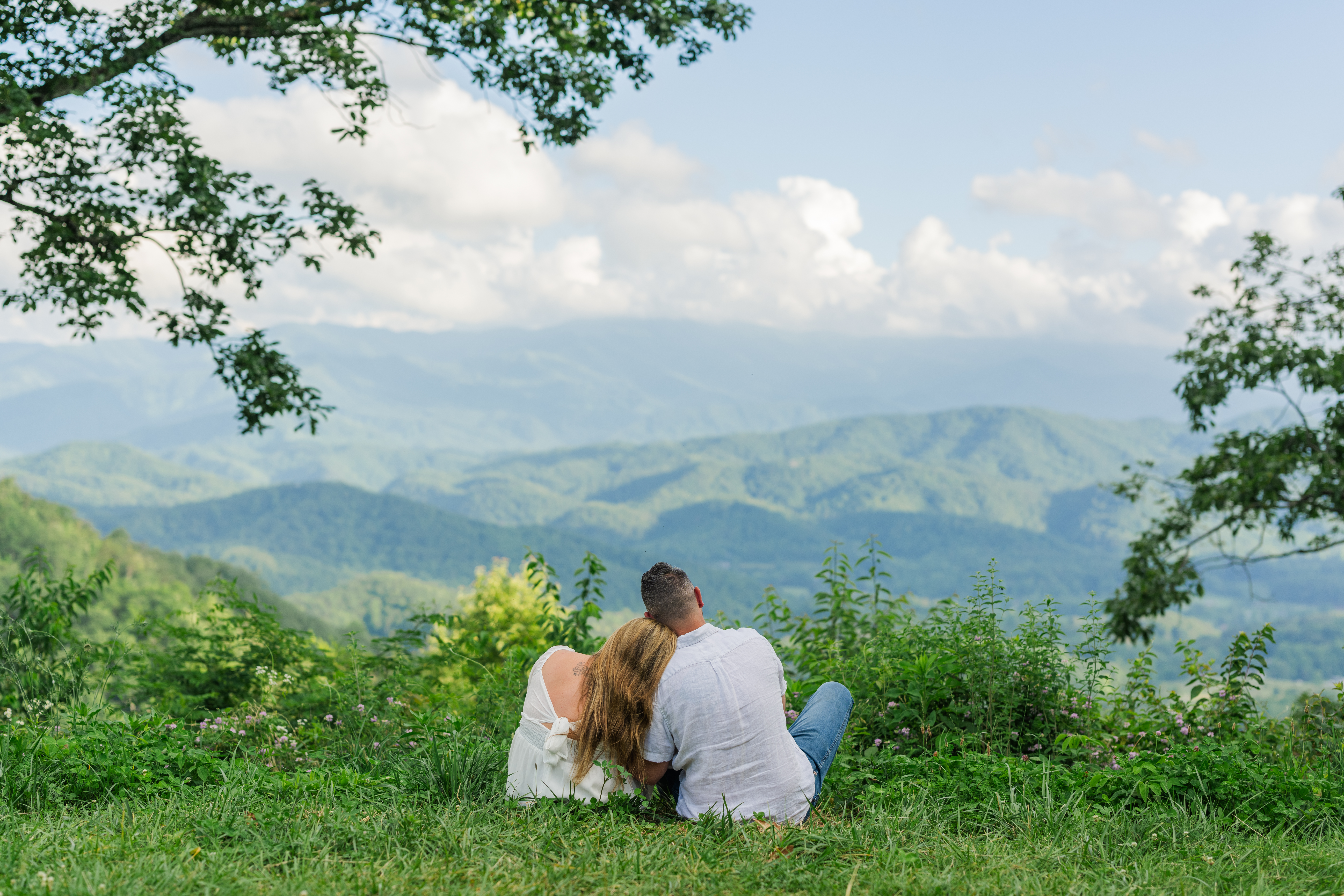Engaged couple sitting cross-legged near cliff overlooking mountains during Smoky Mountains engagement session in Townsend, Tennessee