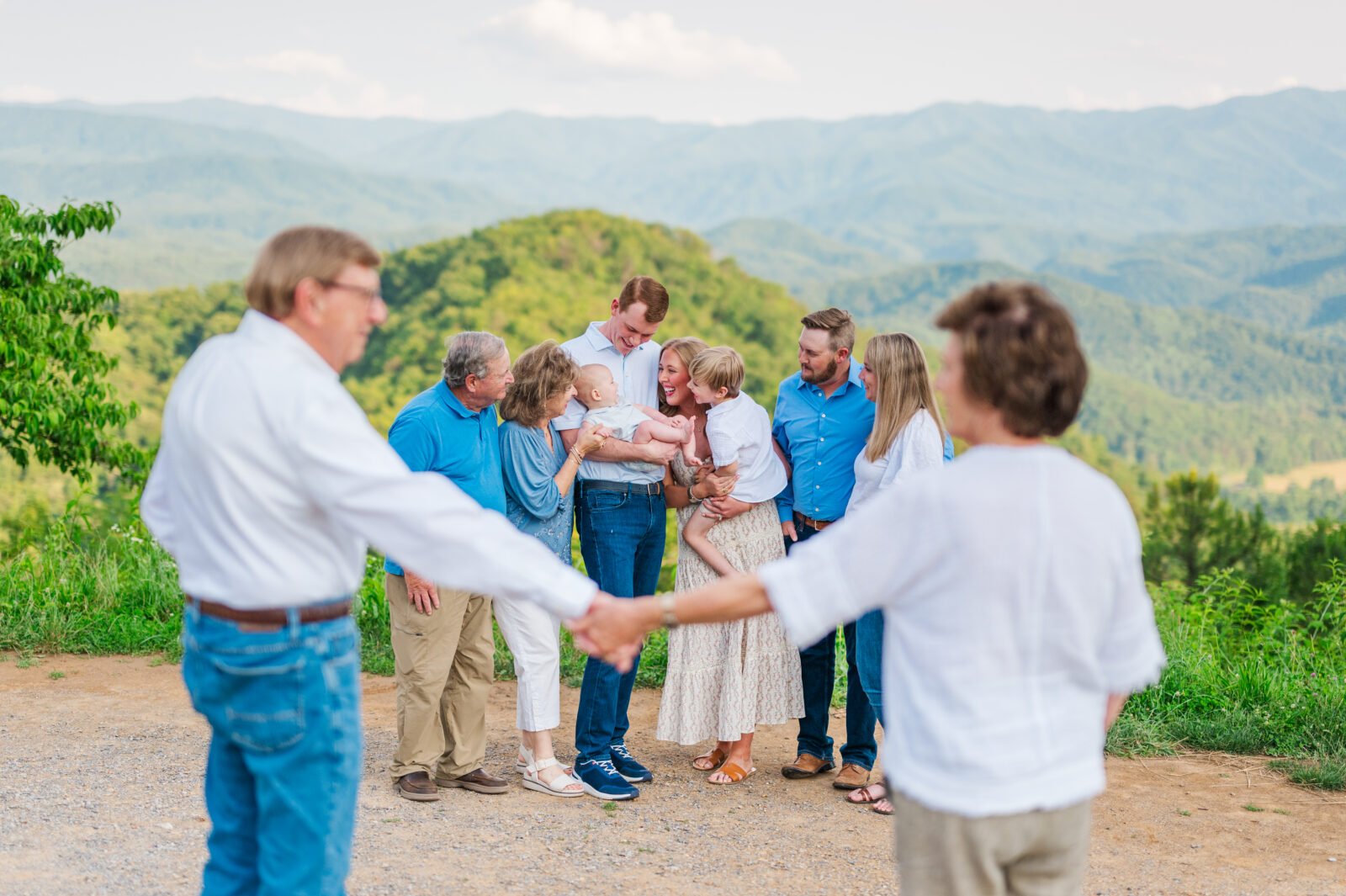 Grandparents hold hands and smile at each other while their children, grandchildren, and great grandchildren play in the distance on vacation in the Smoky Mountains during a family portrait session in Gatlinburg