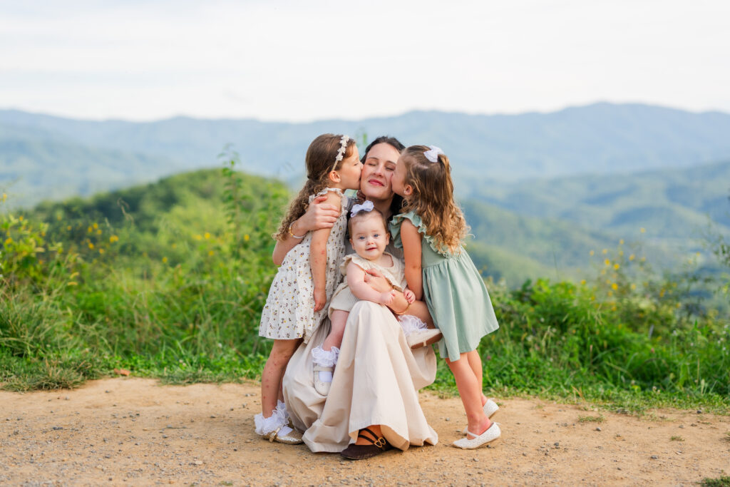A photographer captures two daughters kissing their mom on the cheek with a baby inter lap smiling at the camera in the Great Smoky Mountains