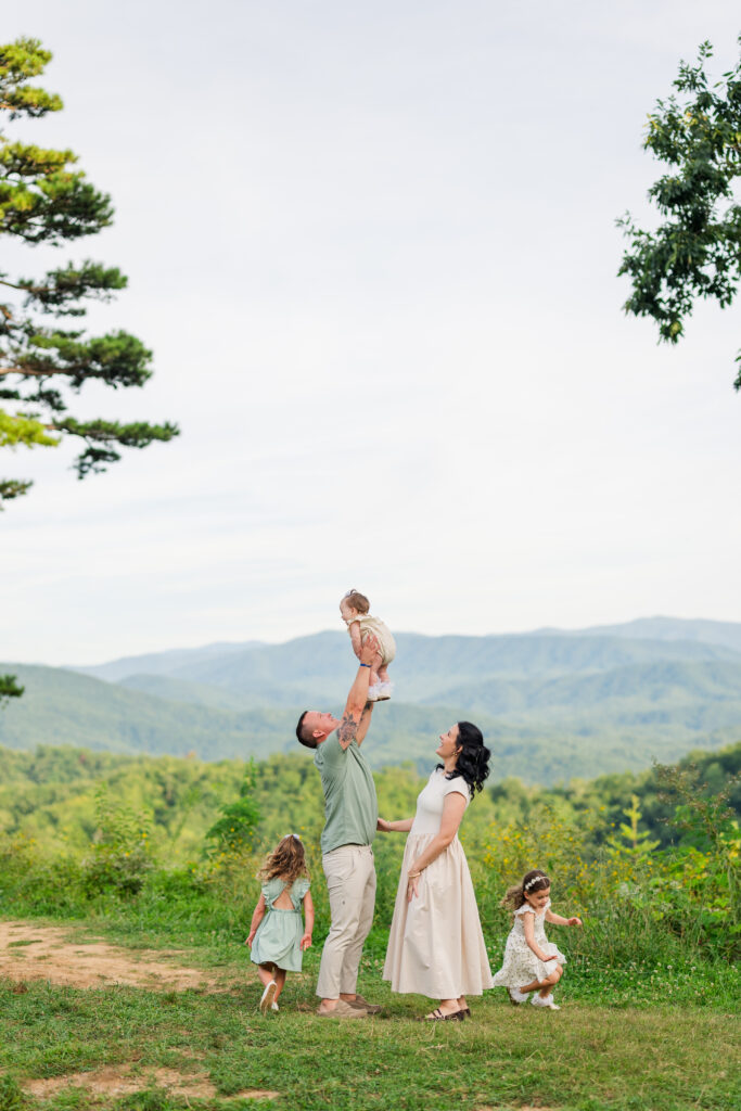 Gatlinburg Photographer captures family portraits of a family on vacation in the Smoky Mountains as the father tosses the baby up with mom looking up and smiling and their two young daughters play tag around them with the mountains in the background in late summer.