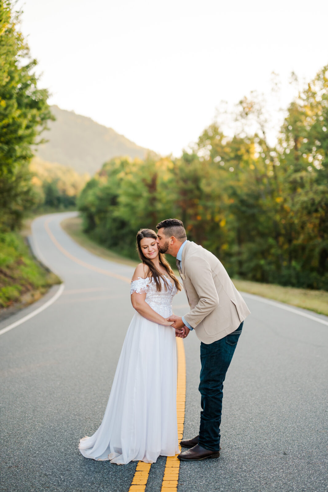 Groom kissing his bride’s cheek on a quiet mountain road in the Smoky Mountains at sunrise — candid Gatlinburg wedding photographer moment.