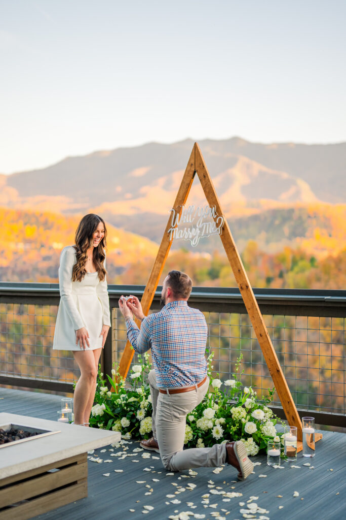 A grand proposal at a cabin balcony in Gatlinburg, Tennessee overlooking mount Leconte in the great smoky mountains national park with a triangle arbor, a neon marry me sign, and beautiful flowers and candles around it