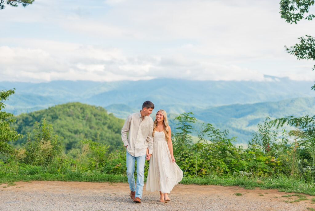 couple holding hands walking with mountain in the background in Gatlinburg Tennessee while a photographer takes their engagement photos
